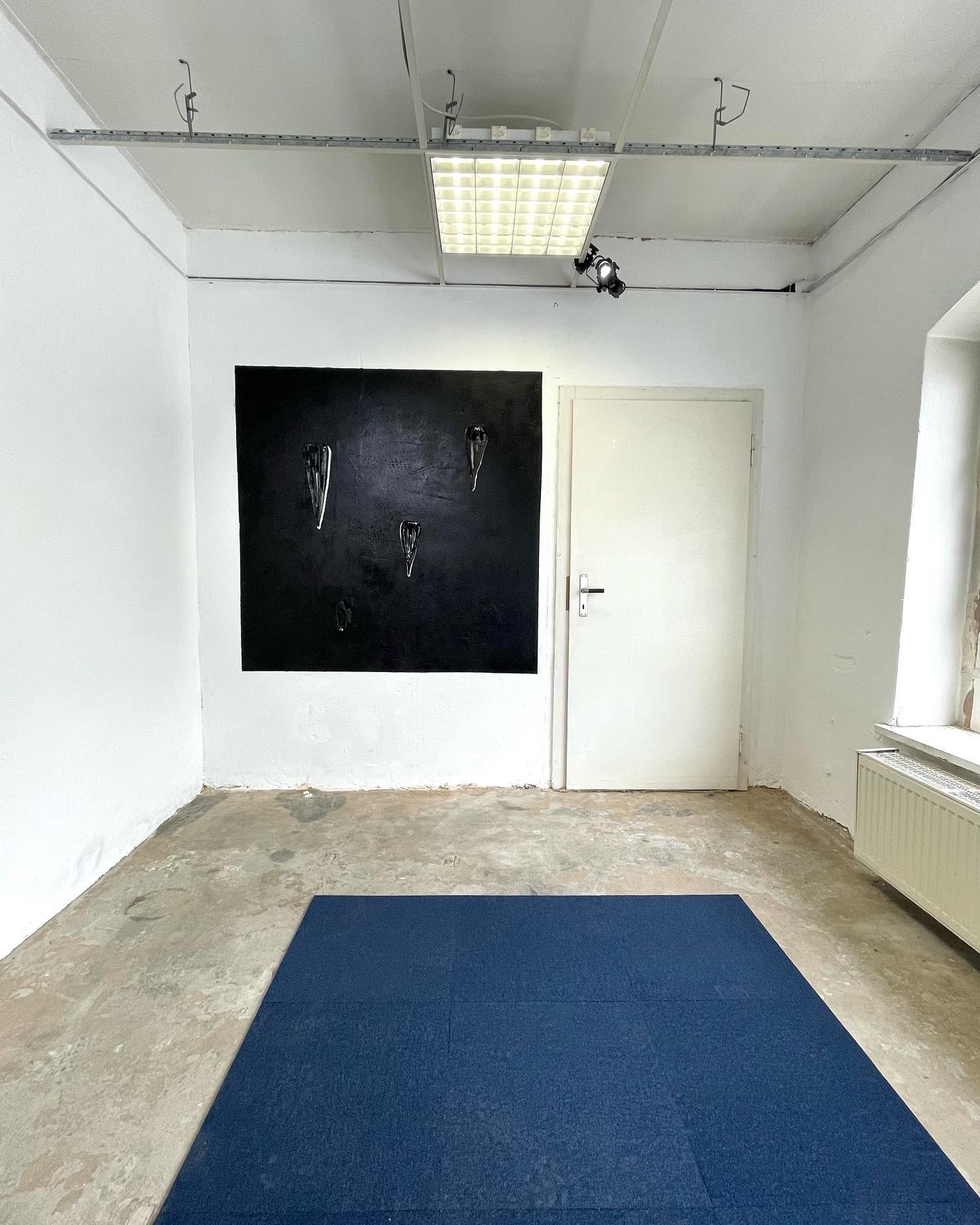 2022년 독일 예나에서 전시한 전시장 전경 이미지이다. 한쪽 벽이 검은색 네모로 칠해져 있고 네모 안쪽으로 세개의 헤어핀 모양의 도자 작업들이 놓여져 있다. 벽고 연결된 바닥에는 벽의 네모와 같은 크기의 파란색 도톰한 매트가 깔려 있다. 벽의 네모 옆으로 흰색의 벽과 다른 아이보리 색상의 문이 있다.