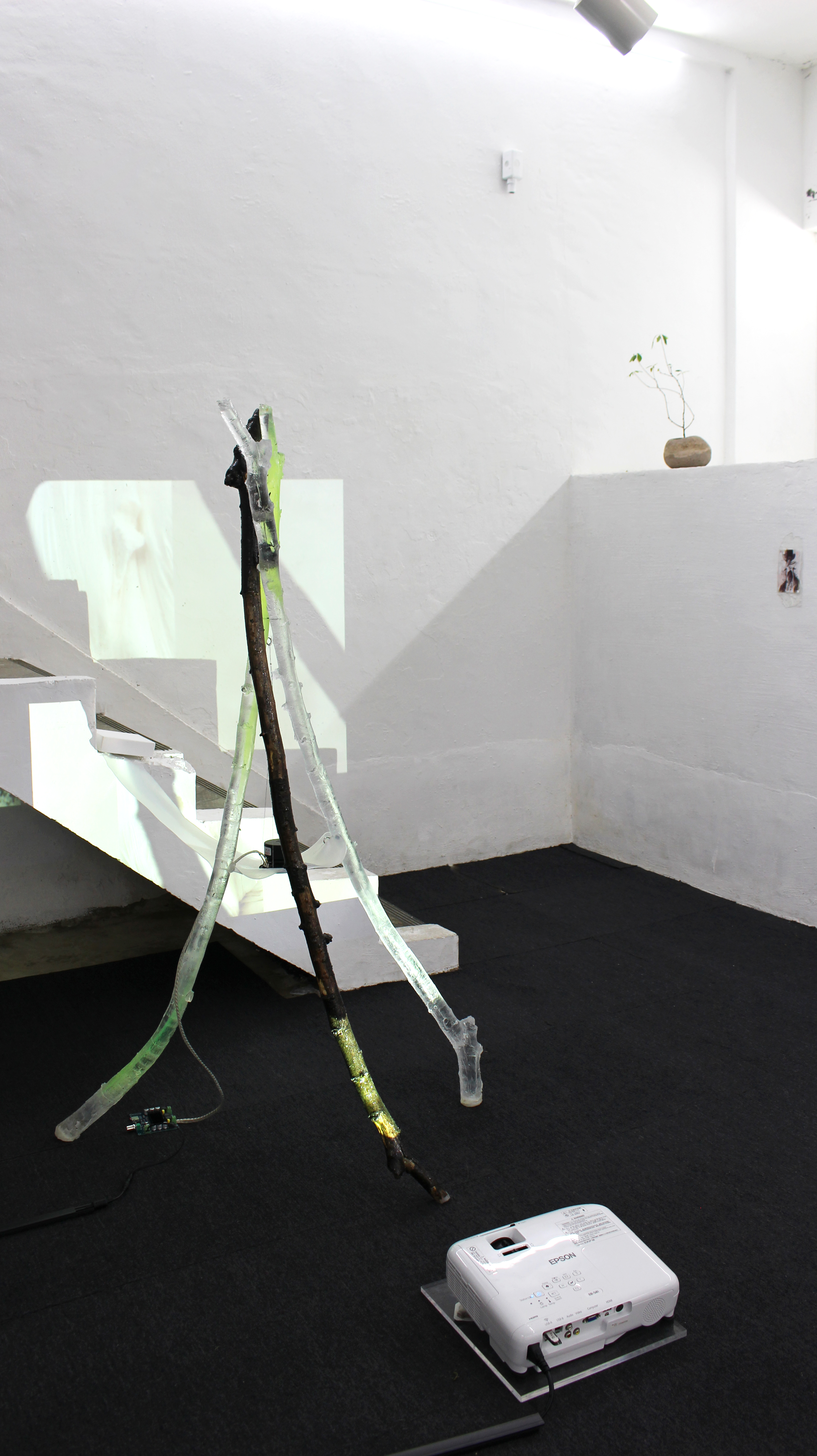 TAT 전시 이미지, 나무 막대와 레진 막대 2개가 3개의 다리가 되어 서있는 작업이 있다. 세 막대의 중심에는 길고 얇은 실리콘 놓여져 있으며 그 위에 스피커가 놓여져 있다. 작품 뒤로 가동되는 프로젝터로 인해 영상은 계단과 일부가 가려진채 보여진다.